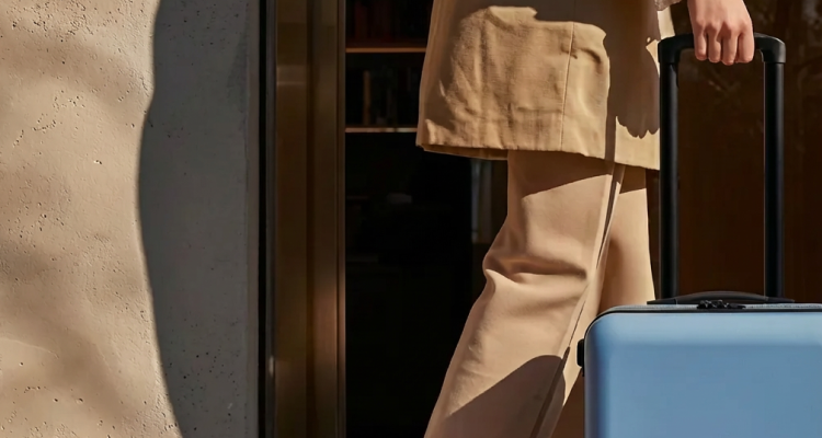 Close-up of a person in beige clothing pulling a blue rolling suitcase through a doorway.