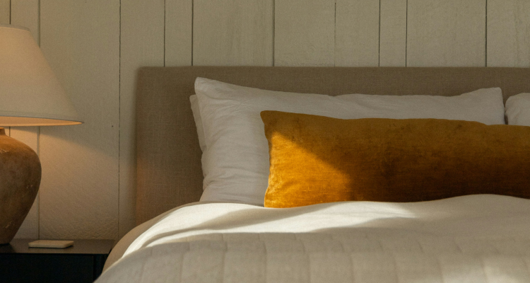 Close-up of a bed with a tan headboard, white duvet, and a long mustard-yellow velvet bolster pillow, lit by a lamp and sunlight.