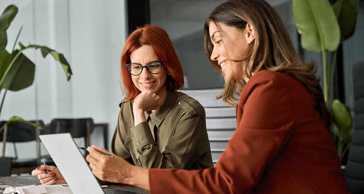 Two professional women smiling while looking at a laptop screen together in a modern office setting